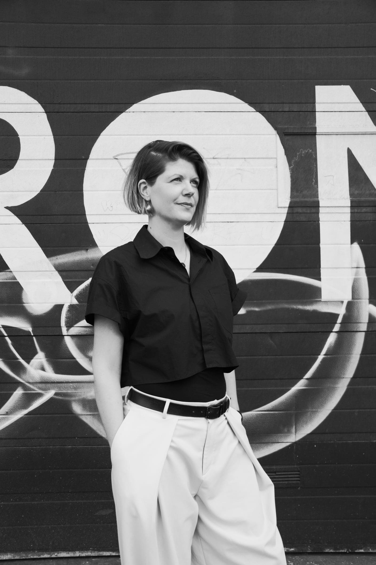Els staat met de handen in de zakken voor een graffitimuur en kijkt opzij